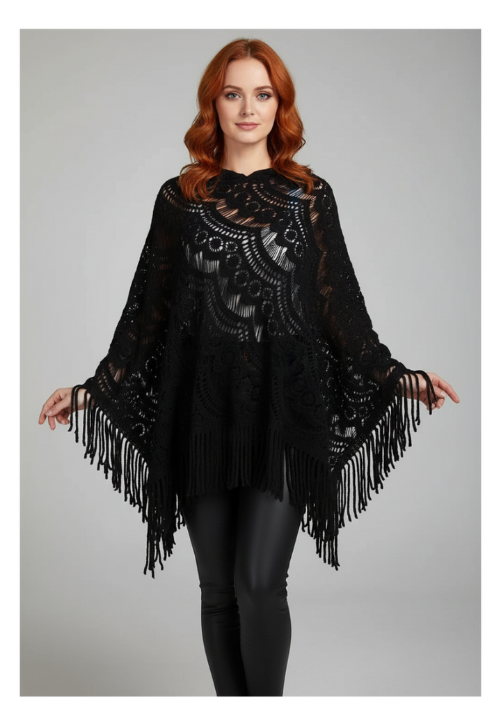 A woman with red hair wearing a black lace and fringe poncho sweater over black leggings, standing against a light gray studio background.